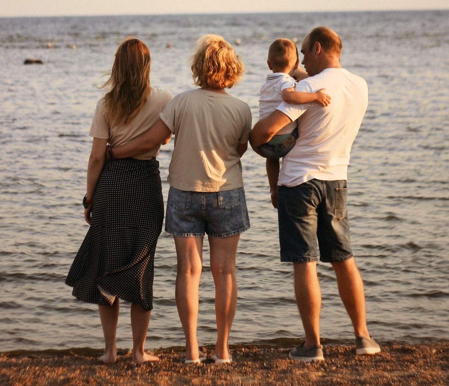 A family with a toddler standing together at the water's edge at golden hour